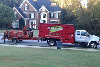 A red truck and wood chipper from Northside Tree company are parked on a suburban street in front of a large brick house with white trim and a well-kept lawn.
