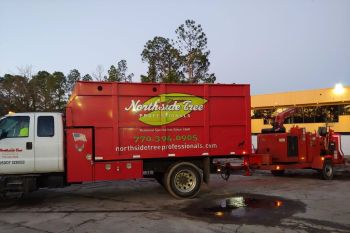A large red Northside Tree Professionals truck and equipment trailer are parked on a concrete lot near trees and a commercial building at dusk. The company’s logo and contact info are visible on the side of the truck.