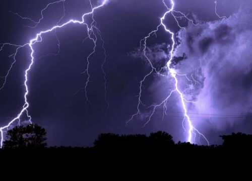 Bright lightning bolts illuminate a dark, cloudy night sky above a silhouetted treeline, with intense flashes branching across the scene.