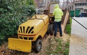 A worker in a yellow hoodie operates a wood chipper, feeding branches into the machine on a residential driveway with houses and trash bins visible in the background.