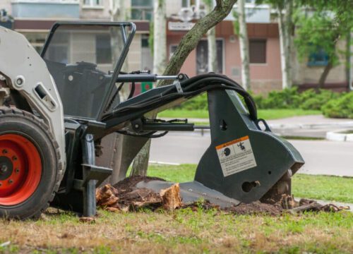 A stump grinder machine attached to a loader is removing a tree stump on a grassy area near a sidewalk and residential buildings.