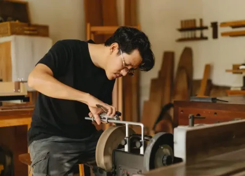 A person wearing glasses and a black shirt is sharpening a tool on a grinding machine in a woodworking shop, surrounded by wooden boards and workshop equipment.