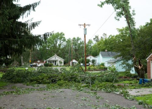 Fallen tree branches and debris block a residential street, with downed power lines and damaged trees visible. Houses and garages are in the background on a cloudy day.