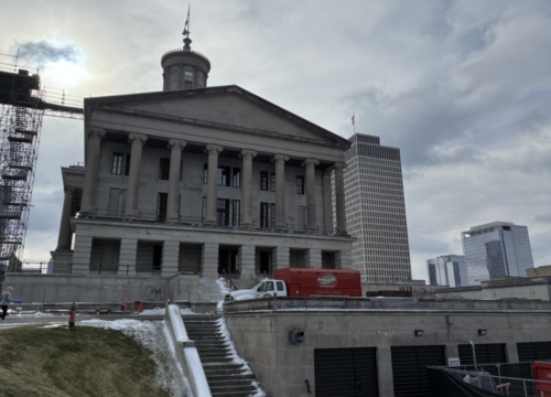 A neoclassical government building with columns and a dome stands on a snowy hill. A red truck is parked nearby, and modern skyscrapers rise in the background under a cloudy sky.