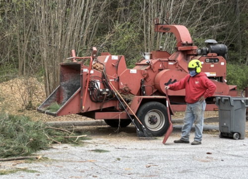 A worker in safety gear stands next to a large red wood chipper on a paved surface, with branches and debris scattered around. Trees and shrubs are in the background.