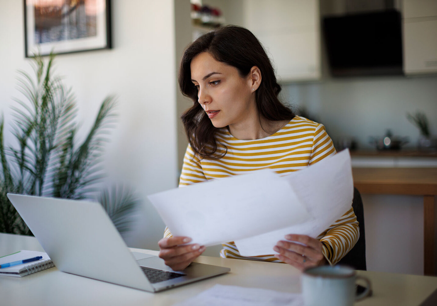 A woman in a yellow striped shirt sits at a table, looking at documents in her hand while working on a laptop. A notebook, pen, and mug are also on the table, with a plant in the background.